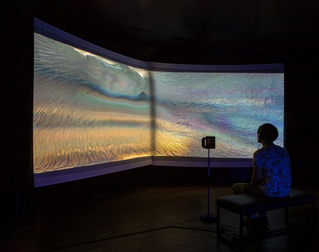 A black silhouette of an individual sits in front of a projected heavily textured background. The combination of the colours resemble the pool of rainbow colours following an oil spill. The iridescence illumination merges from the centre of the screen bursting outwards in asynchronous waves. In front of the seated individual is an ipad in their periphery on a stand. Credit: Augmented Body, Altered Mind by AlanJames Burns